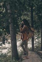 cattura il Visualizza. pieno lunghezza di giovane moderno donna con zaino Fotografare natura mentre escursioni a piedi nel il boschi foto