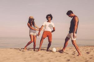 appena avendo divertimento. tre allegro giovane persone giocando con calcio palla su il spiaggia con mare nel il sfondo foto