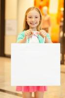 poco ragazza con shopping Borsa. allegro poco ragazza Tenere shopping Borsa e sorridente a telecamera mentre in piedi nel shopping centro commerciale foto