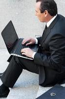 concentrato su opera. fiducioso anziano uomo nel vestito formale Lavorando su il computer portatile mentre seduta all'aperto foto