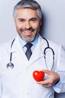fiducioso cardiologo. fiducioso maturo medico Tenere cuore forma giocattolo e sorridente mentre in piedi contro grigio sfondo foto