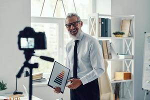 contento maturo uomo nel elegante camicia e cravatta mostrando grafico e condivisione attività commerciale Esperienza mentre fabbricazione sociale media video foto