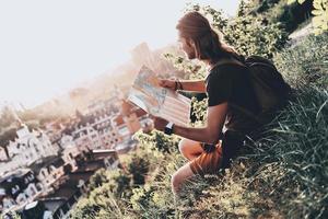 godendo il viaggio. bello giovane uomo nel casuale capi di abbigliamento utilizzando carta geografica mentre seduta su il collina all'aperto foto