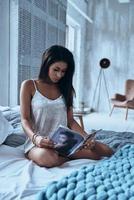 pigro mattina a casa. attraente giovane donna lettura moda rivista mentre seduta su il letto a casa foto