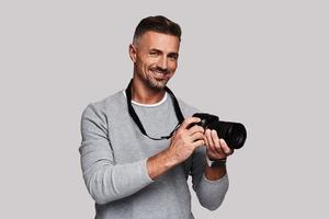 il suo migliore passatempo. bene guardare giovane uomo Tenere digitale telecamera e sorridente mentre in piedi contro grigio sfondo foto