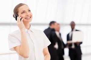 donna d'affari su il Telefono. allegro giovane donna nel bianca camicia parlando su il mobile Telefono e sorridente mentre il suo colleghi in piedi su sfondo foto