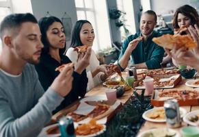 migliore gli amici. gruppo di giovane persone nel casuale indossare mangiare Pizza e sorridente mentre avendo un' cena festa in casa foto