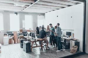 riuscito attività commerciale squadra. pieno lunghezza di giovane moderno persone nel inteligente casuale indossare avendo un' incontro mentre in piedi dietro a il bicchiere parete nel il creativo ufficio foto