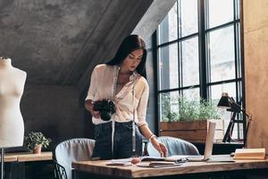 sviluppando nuovo moda linea. concentrato giovane donna con nastro misurare su sua collo guardare a moda rivista e Tenere digitale telecamera mentre in piedi vicino il scrivania nel sua laboratorio foto