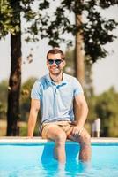 la spesa il suo tempo libero tempo bordo piscina. bello giovane uomo nel occhiali da sole seduta di il piscina e sorridente foto