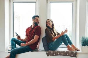 moderno giovane coppia nel casuale capi di abbigliamento bonding insieme e sorridente mentre seduta su il finestra davanzale in casa foto