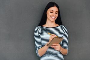 fabbricazione alcuni Appunti. attraente giovane donna scrittura qualcosa nel sua Nota tampone mentre in piedi contro grigio sfondo foto