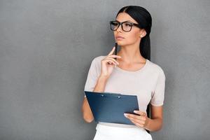 fabbricazione attività commerciale pianificazione. riflessivo giovane donna d'affari Tenere appunti e guardare lontano mentre in piedi contro grigio sfondo foto