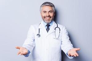 allegro medico. allegro maturo grigio capelli medico guardare a telecamera mentre allungamento su mani e sorridente mentre in piedi contro grigio sfondo foto