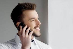 bene parlare. bello giovane uomo nel vestito formale parlando su il mobile Telefono e sorridente foto