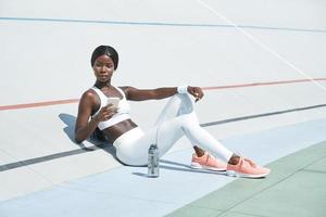 bellissimo giovane africano donna nel gli sport capi di abbigliamento Tenere inteligente Telefono mentre seduta su gli sport traccia all'aperto foto