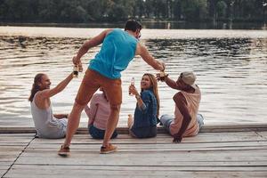 sensazione gratuito per fare qualsiasi cosa. gruppo di contento giovane persone nel casuale indossare sorridente e potabile birra mentre seduta su il molo foto