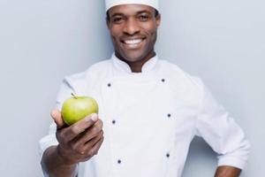 mangiare un' salutare pasto contento giovane africano capocuoco nel bianca uniforme allungamento su verde Mela e guardare a telecamera con Sorridi mentre in piedi contro grigio sfondo foto