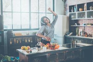 godendo sua mattina. bellissimo giovane misto gara donna nel cuffie cucinando insalata e danza mentre in piedi nel cucina a casa foto