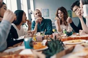 grande cibo e grande le persone. gruppo di giovane persone nel casuale indossare mangiare Pizza e sorridente mentre avendo un' cena festa in casa foto