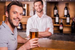 contento bar cliente. contento bar cliente Tenere bicchiere di birra e sorridente mentre barista in piedi nel il sfondo foto