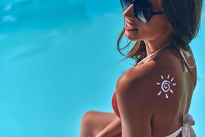 assunzione cura di sua pelle. attraente giovane donna con un' disegnato sole su sua spalla prendere il sole mentre seduta di il piscina all'aperto foto