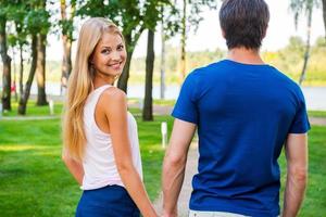 sensazione fiducioso e sicuro vicino lui. posteriore Visualizza di bellissimo giovane donna guardare al di sopra di spalla e sorridente mentre a piedi lungo il parco strada con sua fidanzato foto