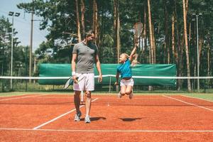 poco vincitore. pieno lunghezza di poco biondo capelli ragazza nel gli sport capi di abbigliamento trasporto tennis racchetta e guardare a sua padre a piedi vicino sua di tennis Tribunale foto