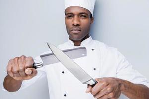 pronto per opera. fiducioso giovane africano capocuoco nel bianca uniforme Tenere coltelli nel il suo mani e guardare a telecamera mentre in piedi contro grigio sfondo foto