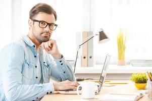 la creazione di nuovo visione. bello giovane uomo nel camicia e occhiali Lavorando su il computer portatile e guardare a telecamera mentre seduta a il suo Lavorando posto foto