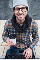 la spesa bene tempo all'aperto. superiore Visualizza di bello giovane uomo Tenere un' tazza di caffè e Lavorando su il computer portatile mentre seduta all'aperto foto