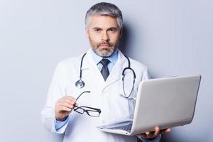 fiducioso e esperto medico. fiducioso maturo medico Lavorando Tenere il computer portatile e guardare a telecamera mentre in piedi contro grigio sfondo foto