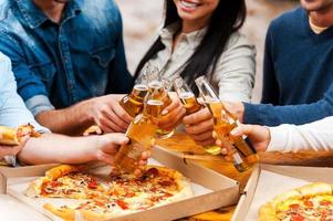 festeggiare loro amicizia. avvicinamento di gruppo di giovane persone mangiare Pizza e tintinnante bicchieri con birra mentre in piedi all'aperto foto