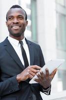 uomo d'affari con digitale tavoletta. fiducioso giovane africano uomo nel vestito formale Lavorando su digitale tavoletta e guardare lontano con Sorridi mentre in piedi all'aperto foto