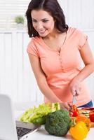 cucinando di ricetta. allegro giovane donna guardare a il il computer portatile e sorridente mentre taglio verdure nel il cucina foto