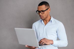 fiducioso esso esperto. fiducioso giovane africano uomo Lavorando su il computer portatile mentre in piedi contro grigio sfondo foto