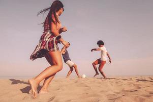 spiaggia volta. tre allegro giovane persone giocando con calcio palla su il spiaggia foto