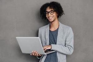 fabbricazione idee accadere. allegro giovane africano uomo Tenere il computer portatile e sorridente mentre in piedi contro grigio sfondo foto