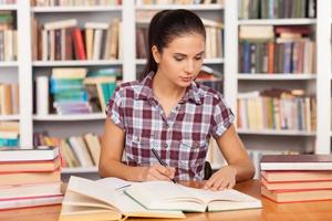 preparazione per il esami. riflessivo giovane donna scrittura qualcosa nel sua Nota tampone e lettura libro mentre seduta a il biblioteca scrivania foto