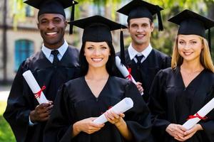 sensazione fiducioso nel loro futuro. quattro Università laureati nel la laurea abiti in piedi vicino per ogni altro e sorridente foto