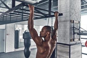 fabbricazione sforzo. A petto nudo giovane africano uomo fare pull-up e guardare lontano mentre esercizio nel il Palestra foto