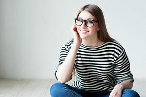 allegro bellezza. bellissimo giovane donna nel a strisce capi di abbigliamento seduta su il legno duro pavimento e Tenere mano su mento foto