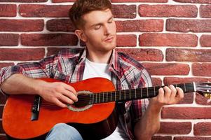 giocando chitarra. bello giovane uomo giocando acustico chitarra mentre pendente a il mattone parete foto