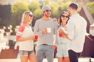 caffè rompere. quattro giovane allegro persone chat e potabile caffè mentre in piedi su il tetto terrazza insieme foto