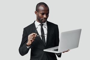 concentrato giovane africano uomo nel vestito formale Lavorando utilizzando computer mentre in piedi contro grigio sfondo foto