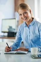 professionale e ambizioso.sorridente giovane donna d'affari nel vestito formale scrittura nel Nota tampone e guardare a telecamera mentre seduta a sua Lavorando posto foto