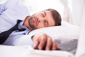 stanco e sovraccarico di lavoro. bello giovane uomo nel camicia e cravatta addormentato nel letto con il suo mano posa su il computer portatile tastiera foto