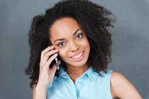 chiamata per voi. ritratto di attraente giovane africano donna parlando su il mobile Telefono e sorridente mentre in piedi contro grigio sfondo foto