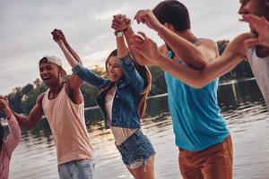 appena avendo divertimento. gruppo di giovane persone nel casuale indossare sorridente e Tenere mani mentre in piedi su il molo foto