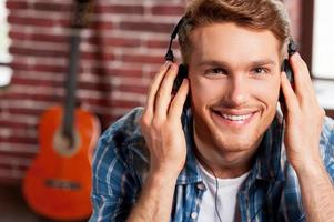 godendo il suo preferito musica. bello giovane uomo regolazione il suo cuffie e sorridente mentre acustico chitarra posa nel il sfondo foto